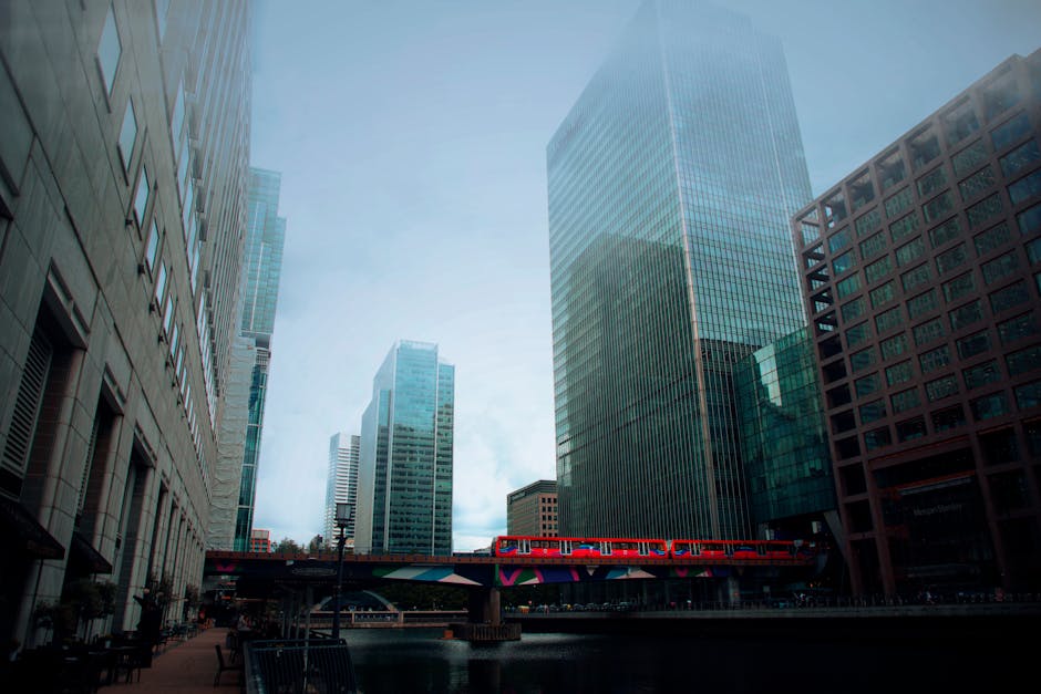 Photograph of a modern cityscape in Canning Town, featuring several tall glass and concrete office buildings and apartment blocks along a waterfront. The buildings vary in height and architectural style, with some reflecting the sky and surrounding structures through their glass facades. In the foreground, a red train is seen crossing a bridge or elevated track, which is decorated with colorful graffiti art. The scene is captured during daylight hours under a slightly overcast sky, with soft, diffused natural lighting. The image portrays an urban environment typical of a busy commercial and residential area, relevant to house removals and moving services provided by Man with Van Canning Town in the context of home relocation or furniture transport amidst city infrastructure.
