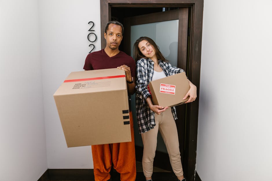 A man and a woman standing inside a residential property near a doorway, both holding cardboard boxes used for packing during a home relocation. The man, dressed in a maroon top and orange trousers, holds a large box with packing labels, while the woman, wearing a denim shirt over a white top and beige trousers, carries a smaller box marked with a 'Fragile' label. The background shows a dark wooden doorframe with the number '200' vertically aligned beside it. The scene takes place on a well-lit indoor corridor, likely part of a professional house removal service, with the manwithvancanningtown.co.uk branding relevant to furniture transport and packing during a moving process. The boxes are made of cardboard, some wrapped in plastic for protection, and the individuals are positioned close to the doorway, ready for loading or unloading. The overall setting reflects typical packing and moving activities involved in an organized household relocation within Canning Town, East London.