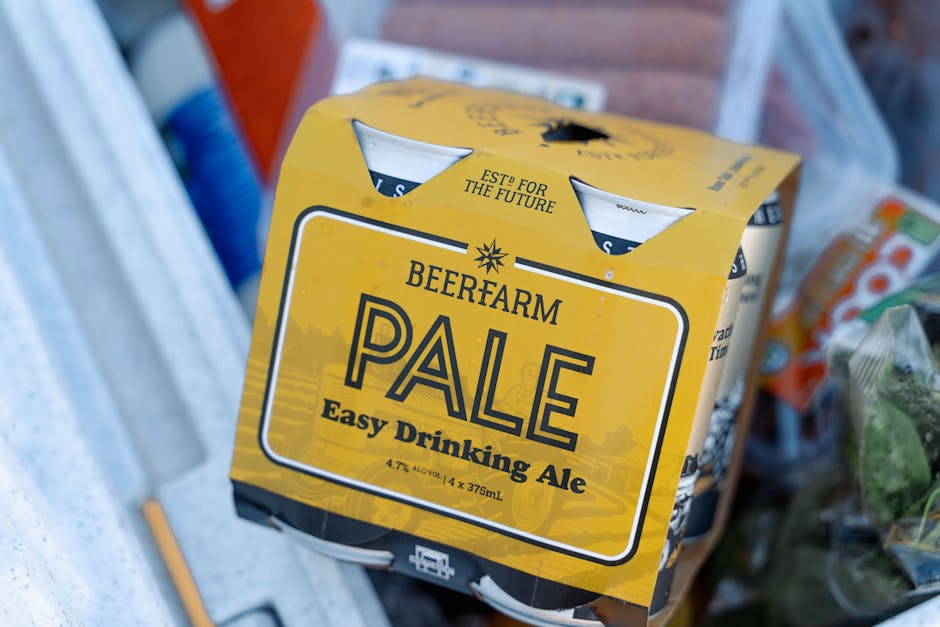 Photograph of a modern cityscape in Canning Town, featuring several tall glass and concrete office buildings and apartment blocks along a waterfront. The buildings vary in height and architectural style, with some reflecting the sky and surrounding structures through their glass facades. In the foreground, a red train is seen crossing a bridge or elevated track, which is decorated with colorful graffiti art. The scene is captured during daylight hours under a slightly overcast sky, with soft, diffused natural lighting. The image portrays an urban environment typical of a busy commercial and residential area, relevant to house removals and moving services provided by Man with Van Canning Town in the context of home relocation or furniture transport amidst city infrastructure.