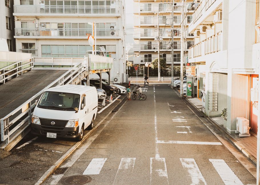 A parking area outside a residential building in Canning Town, featuring a white commercial van parked in one of the designated spaces, which is part of a home relocation or furniture transport process managed by Man with Van Canning Town. The scene includes a row of parked cars, a bicycle being ridden by a cyclist in the background, and a pedestrian walking along the pavement. To the left, a facility with a ramp and metal railings is visible, suggesting accessible loading arrangements for moving furniture and boxes. The pavement shows white painted lines indicating parking bays and pedestrian crossings, with some boxes and packing materials possibly prepared for the move. The background displays modern apartment buildings with balconies, and a green roadside parking meter is seen near the building entrance. The overall lighting is natural, with soft sunlight illuminating the scene, reflecting typical urban moving logistics where loading, unloading, and parking permits might be relevant, which aligns with the considerations around parking permits for a moving van in Canning Town.