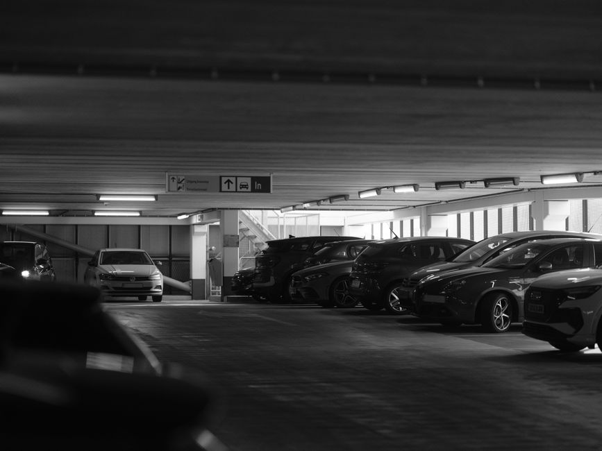 A parking area outside a residential building in Canning Town, featuring a white commercial van parked in one of the designated spaces, which is part of a home relocation or furniture transport process managed by Man with Van Canning Town. The scene includes a row of parked cars, a bicycle being ridden by a cyclist in the background, and a pedestrian walking along the pavement. To the left, a facility with a ramp and metal railings is visible, suggesting accessible loading arrangements for moving furniture and boxes. The pavement shows white painted lines indicating parking bays and pedestrian crossings, with some boxes and packing materials possibly prepared for the move. The background displays modern apartment buildings with balconies, and a green roadside parking meter is seen near the building entrance. The overall lighting is natural, with soft sunlight illuminating the scene, reflecting typical urban moving logistics where loading, unloading, and parking permits might be relevant, which aligns with the considerations around parking permits for a moving van in Canning Town.