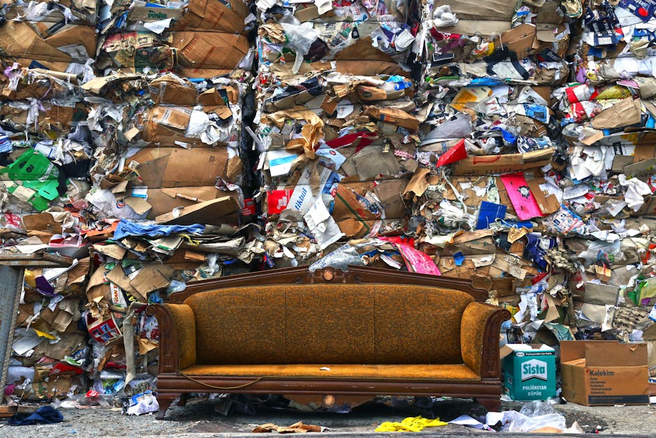 A vintage upholstered sofa with wooden frame and floral fabric sits on a pavement in front of a large pile of mixed household waste, including crumpled cardboard boxes, paper packaging, plastic wrappers, and discarded packaging materials. The waste is stacked high against a wall, with some items spilling onto the ground nearby, and the sofa appears to be part of a bulky waste collection or removal process. The scene is outdoors, with natural lighting illuminating the area, and the setting suggests a house clearance or a bulk waste collection in progress. This image reflects the type of items handled during home relocation and furniture transport services offered by Man with Van Canning Town, particularly in the context of E16 bulky waste removals such as sofas, mattresses, and white goods.
