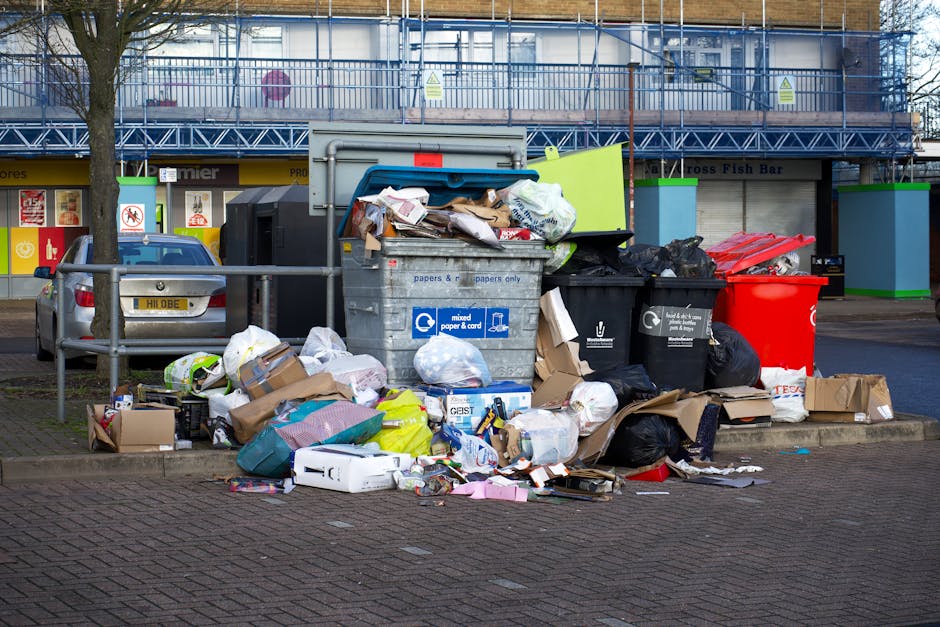An outdoor scene depicting a large accumulation of mixed waste and bulky rubbish in a recycling area near a commercial building, consisting of overflowing cardboard boxes, plastic bags, paper waste, and packaging materials, some spilling onto the paved ground. Multiple wheelie bins, including black, red, and black with labels for paper and general waste, are positioned behind the pile. A grey waste container with a partially open lid is visible, filled with flattened cardboard and other refuse. To the left, a silver car is parked, and a metal railing separates the waste area from the parking spots. The background features a multi-storey building under construction, covered with scaffolding and protective sheeting, with shop signs for local businesses. The scene is lit by daylight, with some shadows cast on the ground, highlighting the mess and disorganization typical of home or commercial waste collection points, as part of a moving and clearance process handled by Man with Van Canning Town.