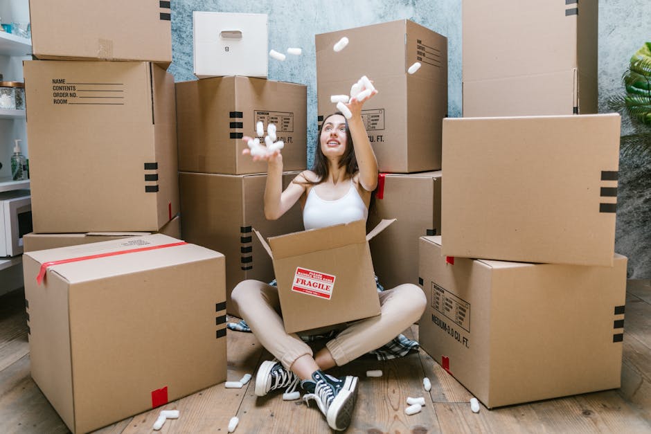A woman sitting on the wooden floor inside a property surrounded by numerous cardboard moving boxes of various sizes, some sealed with red tape and others open, with packing materials like packing peanuts in her hands. The boxes are stacked against a textured grey wall, with a doorway visible behind her. She appears to be mid-action, tossing packing peanuts into the air, with a cheerful expression on her face. The scene also includes items such as a small white microwave on a nearby surface and a green plant partially visible on the right side. The environment is well-lit, indicating natural or artificial light conducive to packing and home relocation activities, as part of a furniture transport or packing and moving process handled by [COMPANY_NAME], supporting residential removals near Canning Town station. This image reflects the packing stage involved in house removals, emphasizing careful handling of belongings during the loading process for a smooth home relocation.