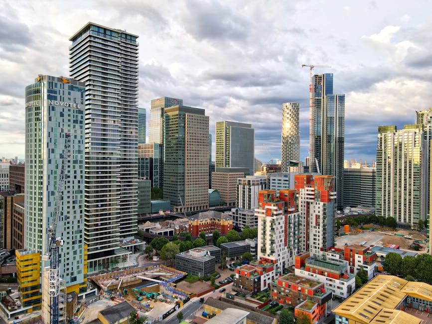 A wide-angle view of a dense urban cityscape featuring numerous high-rise buildings with modern glass facades, some under construction with visible cranes. In the foreground, there are smaller residential and commercial structures, including red brick buildings and white apartment blocks with balconies. The scene is illuminated by natural daylight with partly cloudy skies overhead, and streets below are lined with trees and vehicles. The image, associated with Man with Van Canning Town's house removals service, reflects a busy city environment suitable for planning home relocations, including the logistics of furniture transport and moving logistics.