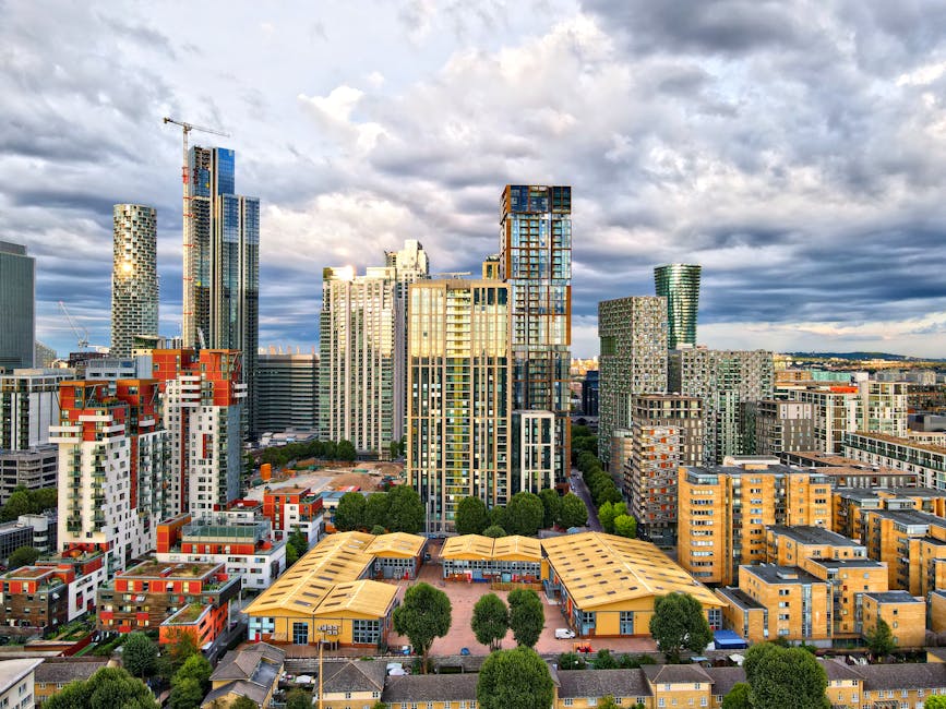 A wide-angle view of a dense urban cityscape featuring numerous high-rise buildings with modern glass facades, some under construction with visible cranes. In the foreground, there are smaller residential and commercial structures, including red brick buildings and white apartment blocks with balconies. The scene is illuminated by natural daylight with partly cloudy skies overhead, and streets below are lined with trees and vehicles. The image, associated with Man with Van Canning Town's house removals service, reflects a busy city environment suitable for planning home relocations, including the logistics of furniture transport and moving logistics.