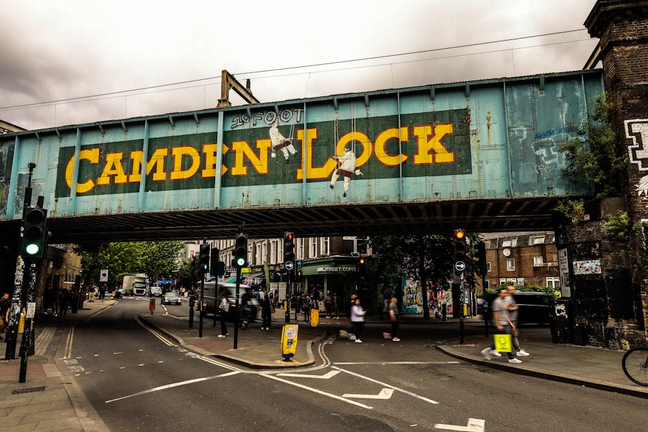 A street scene in Canning Town featuring an old railway bridge with a large, weathered turquoise metal structure that displays the 'Camden Lock' sign painted in yellow letters. The bridge has graffiti and artwork, including illustrations of children hanging from the structure, with one child holding a ball and another climbing a rope. Below the bridge, the busy pavement and road are visible, with pedestrians walking along the sidewalk, some carrying bags. Traffic lights and street signs line the intersection, with cars and delivery trucks moving through the area. The environment indicates an urban setting with a mix of commercial and residential buildings, and greenery visible around the area. This scene depicts an active home relocation or moving logistics environment, where furniture and boxes might be transported through this busy street, supported by local moving services like Man with Van Canning Town.