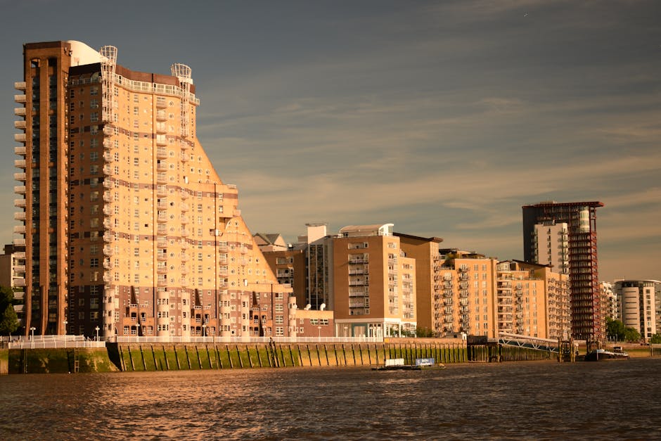 A row of modern multi-storey residential buildings situated along the waterfront in Canning Town, with some structures featuring curved facades and extensive glass windows. The buildings are primarily constructed with brick and concrete, with balconies visible on several levels. The foreground shows the river or canal with calm water, and a concrete or stone embankment running parallel to the water's edge. The sky is partly cloudy, casting subtle shadows on the buildings, indicating late afternoon or early evening lighting. Behind the buildings, the city skyline continues with additional high-rise buildings. There are no visible vehicles, furniture, boxes, or moving equipment in this image, but it establishes a cityscape scene relevant to urban house removals and residential relocations, supporting the context of home relocation services provided by Man with Van Canning Town.