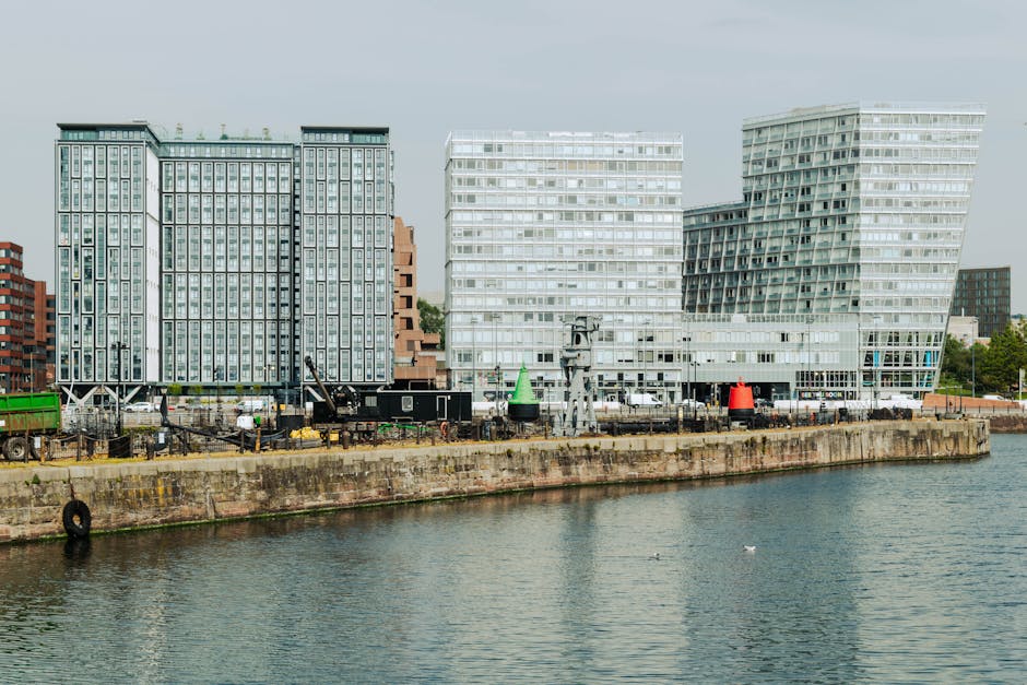 A row of modern multi-storey residential buildings situated along the waterfront in Canning Town, with some structures featuring curved facades and extensive glass windows. The buildings are primarily constructed with brick and concrete, with balconies visible on several levels. The foreground shows the river or canal with calm water, and a concrete or stone embankment running parallel to the water's edge. The sky is partly cloudy, casting subtle shadows on the buildings, indicating late afternoon or early evening lighting. Behind the buildings, the city skyline continues with additional high-rise buildings. There are no visible vehicles, furniture, boxes, or moving equipment in this image, but it establishes a cityscape scene relevant to urban house removals and residential relocations, supporting the context of home relocation services provided by Man with Van Canning Town.