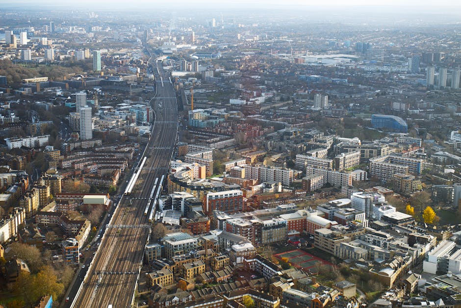 An aerial view of a densely built urban area with numerous residential and commercial buildings, wide roads, and railway tracks stretching through the cityscape. In the foreground, there is a property with a partially open doorway leading inside, where cardboard boxes, wrapped furniture, and packing materials are visible, indicating a home relocation or moving process. A small moving trolley and protective blankets are positioned near the entrance, suggesting the loading or unloading of household items. Outdoors, a man with a van is seen assisting with furniture transport, carefully carrying a large cardboard box. The backdrop includes multi-storey buildings, green spaces, and distant skyscrapers under a clear sky. This scene exemplifies a professional house removals operation within a busy urban environment, aligned with the services offered by Man with Van Canning Town in facilitating local moving routes from Silvertown Estate to Canning Town.