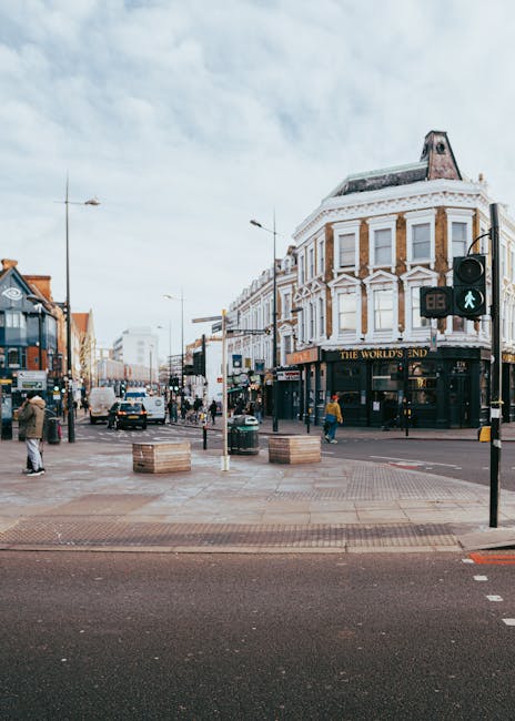 A street scene in Canning Town featuring a pedestrian crossing with a green walking signal, and a Victorian-style building on the corner with a sign reading 'The World's End.' The street is busy with several people walking on the sidewalks, some waiting at the crossing, and vehicles including cars and a double-decker bus are moving along the road. There are lampposts, traffic lights, and street furniture such as planters and benches, with a partly cloudy sky overhead. This setting provides context for house removals and furniture transport, illustrating the urban environment where a professional removal service like Man with Van Canning Town might operate during a home relocation or moving process that involves navigating city streets and navigating loading areas.