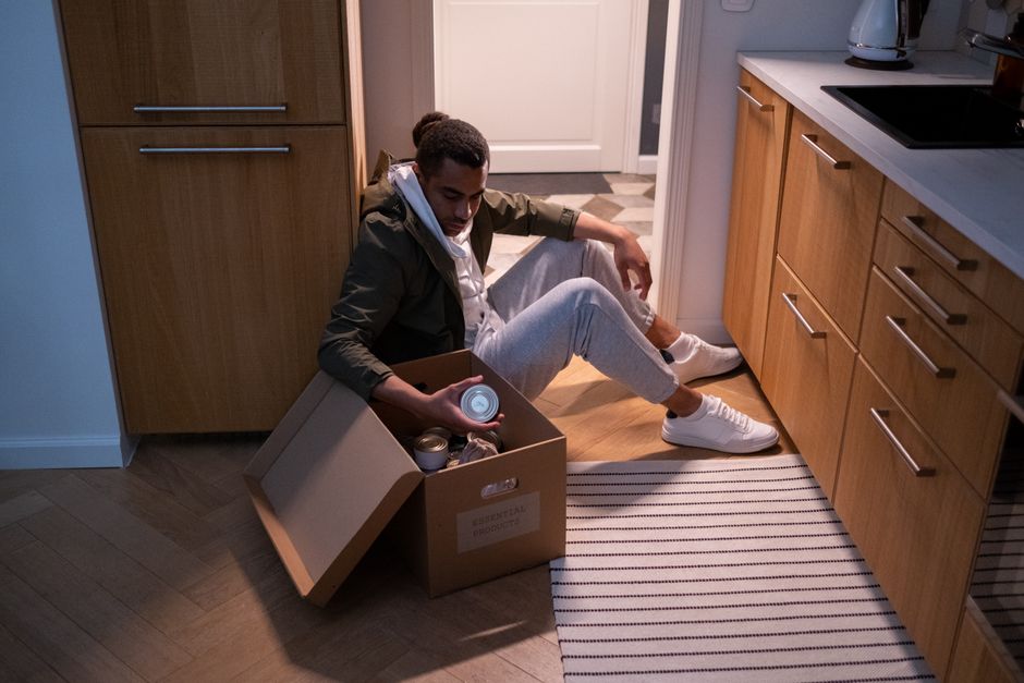 A young man with dark hair and light skin, dressed in a khaki jacket, white hoodie, light grey joggers, and white sneakers, is seated on the wooden floor of a kitchen. He is leaning against a tall wooden cupboard with a neutral expression, holding a small cardboard box filled with various canned food tins and jars. The box, labeled 'Essential Provisions,' rests on the floor beside him, with the lid propped open. The kitchen features wooden cabinets with metallic handles, a white countertop, and a striped rug on the floor. Behind the man, there is a doorway leading to another room, and the scene appears to be part of a home relocation or packing process. The lighting is soft and natural, emphasizing the preparation associated with house removals and furniture transport as part of a professional moving service by Man with Van Canning Town.